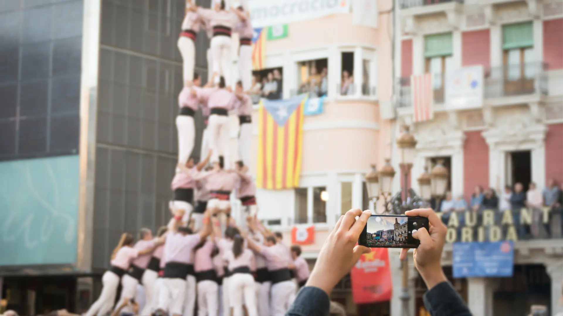 Two hands holding a phone camera and a human tower in the background.