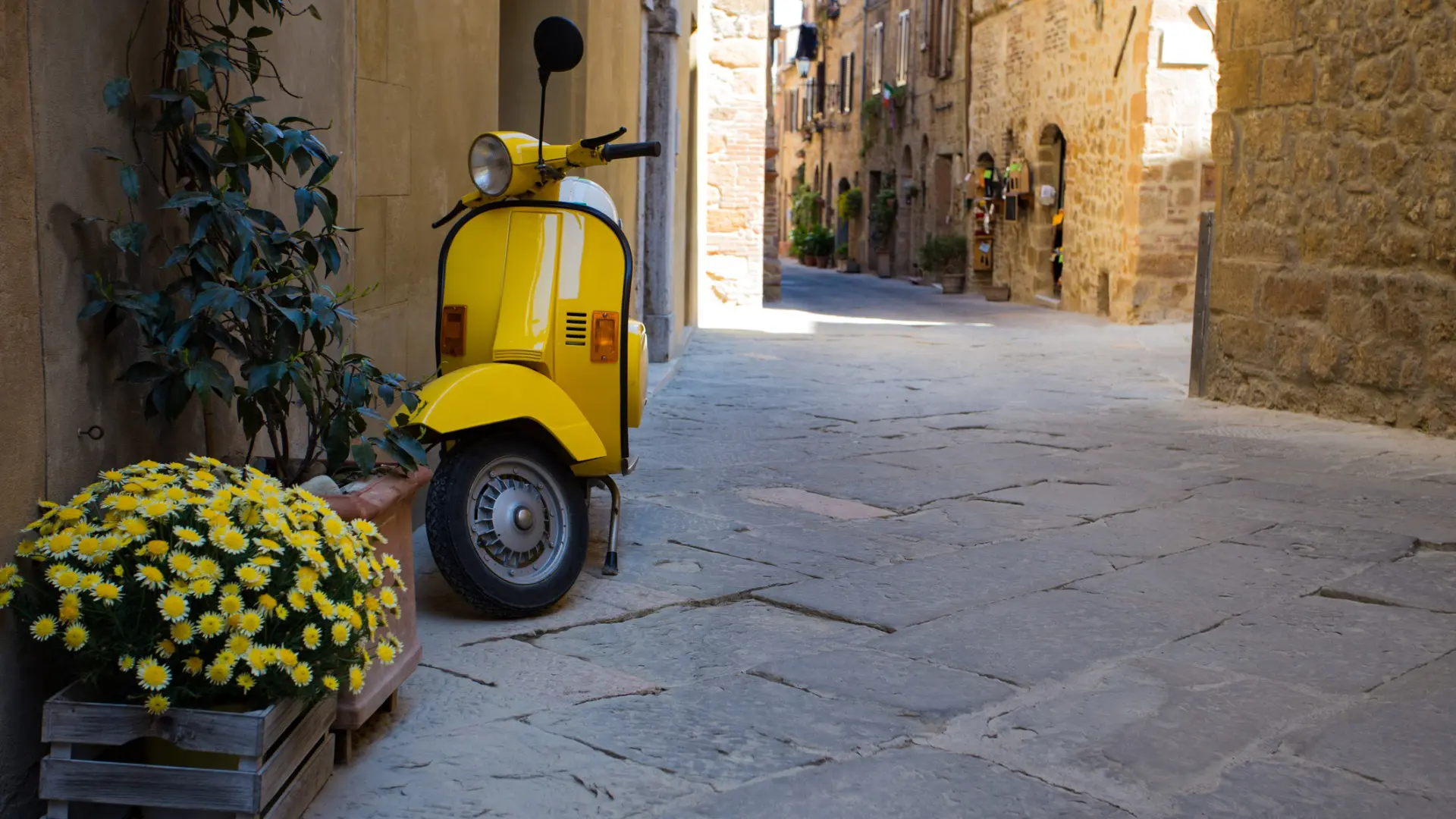 This image shows a yellow vintage vespa parked in a picturesque street in Italy.