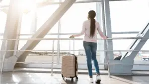 This image shows the rear view of a woman looking at a plane taking off from inside an airport window. She's holding her carry-on suitcase. It's the feature image for our article How to pack for a trip to Europe.
