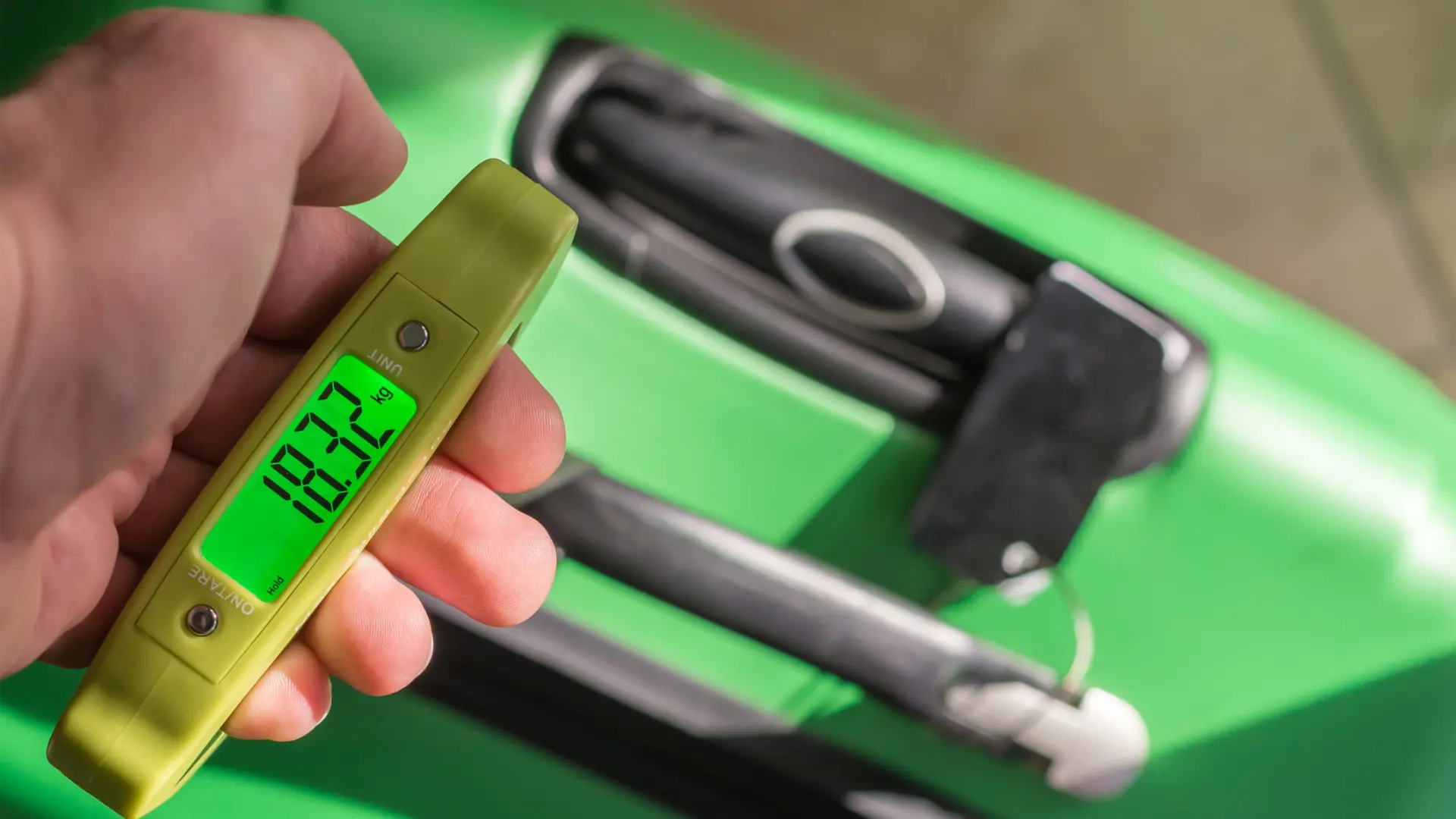 This image shows a hand holding a portable scale, weighing a green suitcase. The scale reads 18.32 kg.