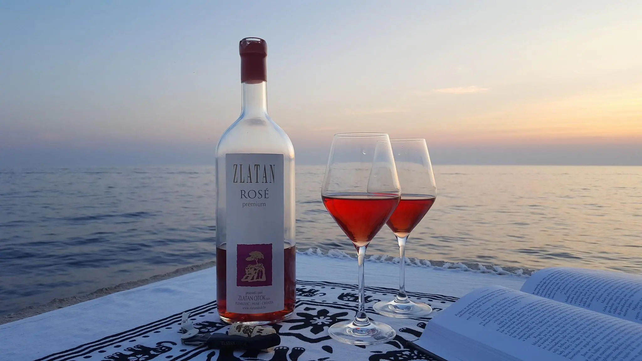 This image shows a half-empty bottle of rose Zlatan Otok wine, two glasses filled with rose wine and an open book with the sea and the sunset in the background.