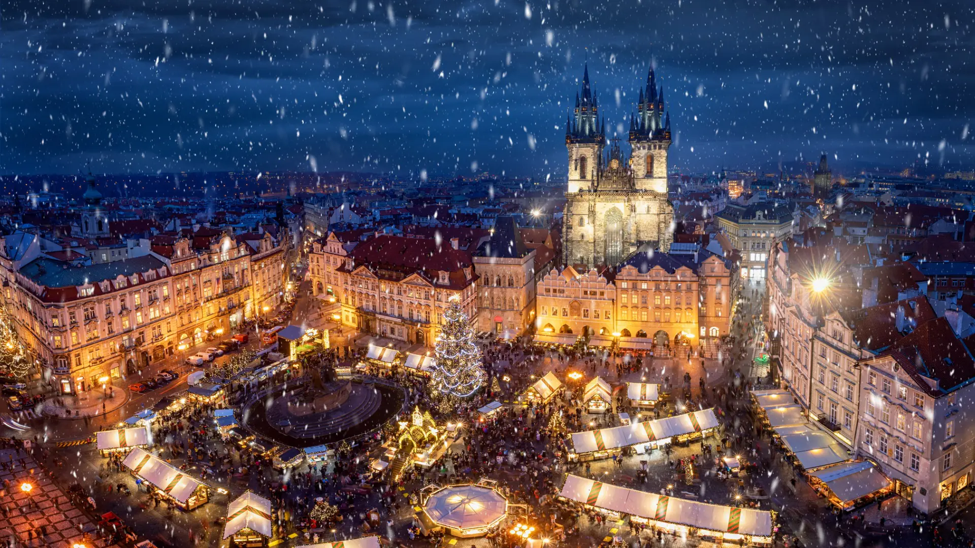 This is an aerial view of the Old Town Square Christmas Market in Prague, packed with people on a snowy evening. 