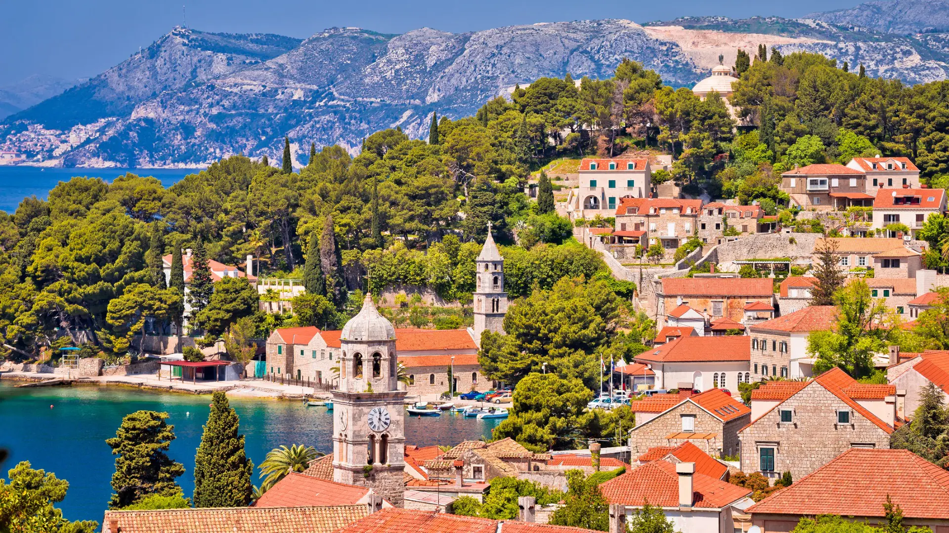 This is a panoramic view of Cavtat. There are stone houses with red-tiled roofs built among trees. In the background, there's a gorgeous beach.