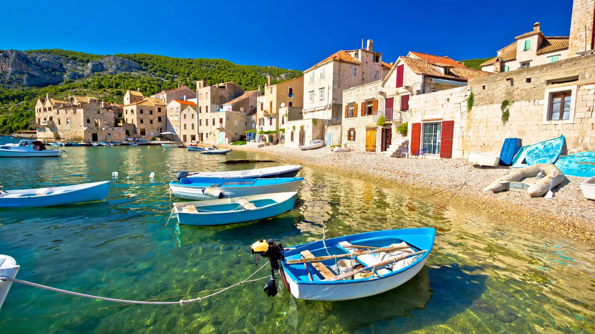 This image shows small wooden fishing boats resting on the crystal clear waters. The shore is lined by traditional stone houses with colorful doors and shutters.