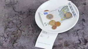 This is a top-down image that shows a small white plate with a 5 EUR bill and some euro coins on a grey table.