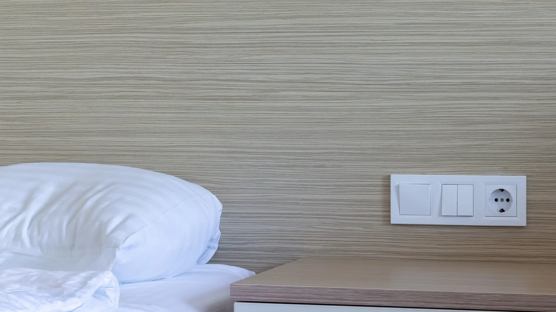 This is a close-up of the sleeping area in a hotel room. There are light switches and a power outlet right next to the pillow and above the bedside table.