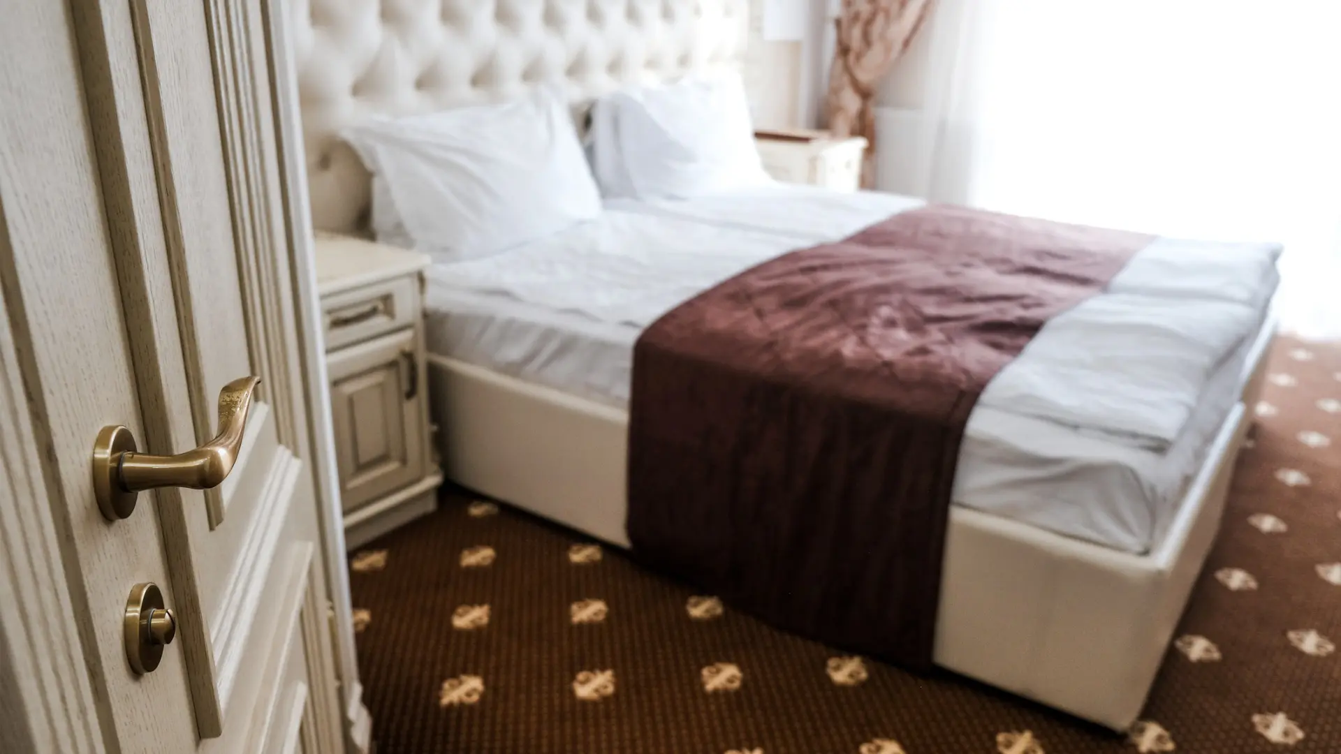 This image shows a hotel room with an old-fashioned bed and bedside table. The floor is covered by carpet from wall to wall.