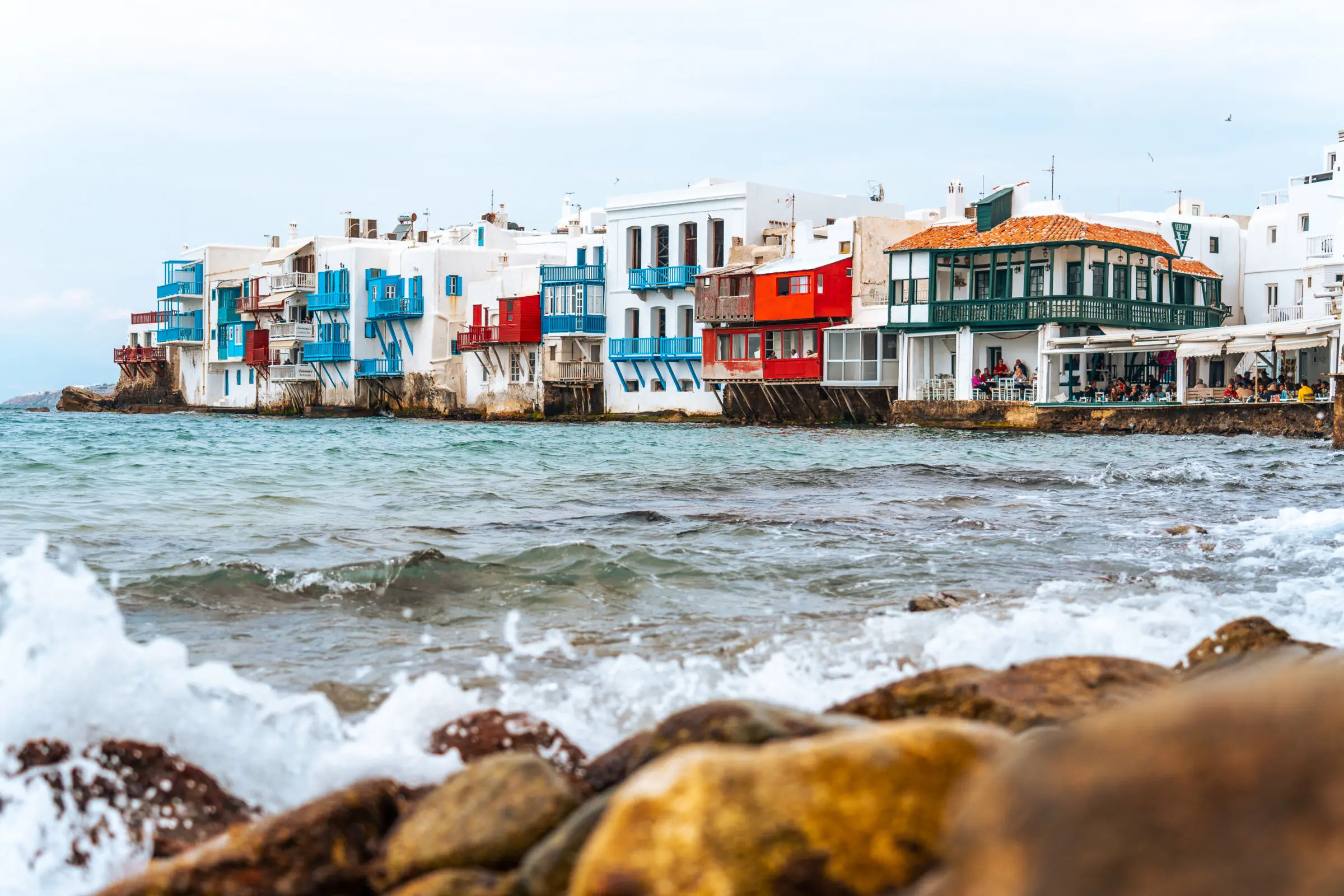 This image shows Little Venice in Mykonos with its characterisitic white houses with colorful wooden balconies built on the edge of the sea.