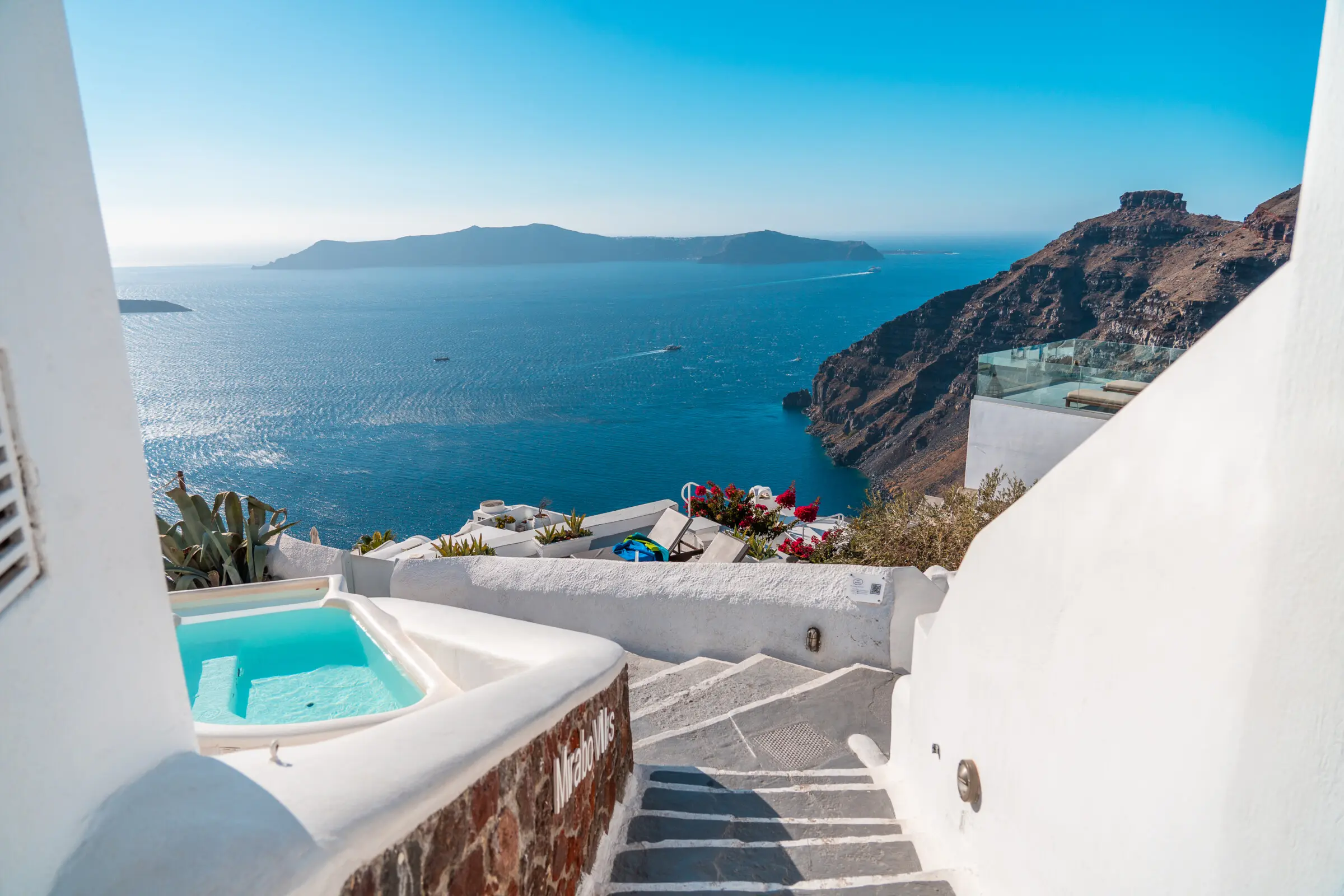 This image shows whitewashed stairs and a small pool in Santorini. The iconic Skaros Rock is in the background.