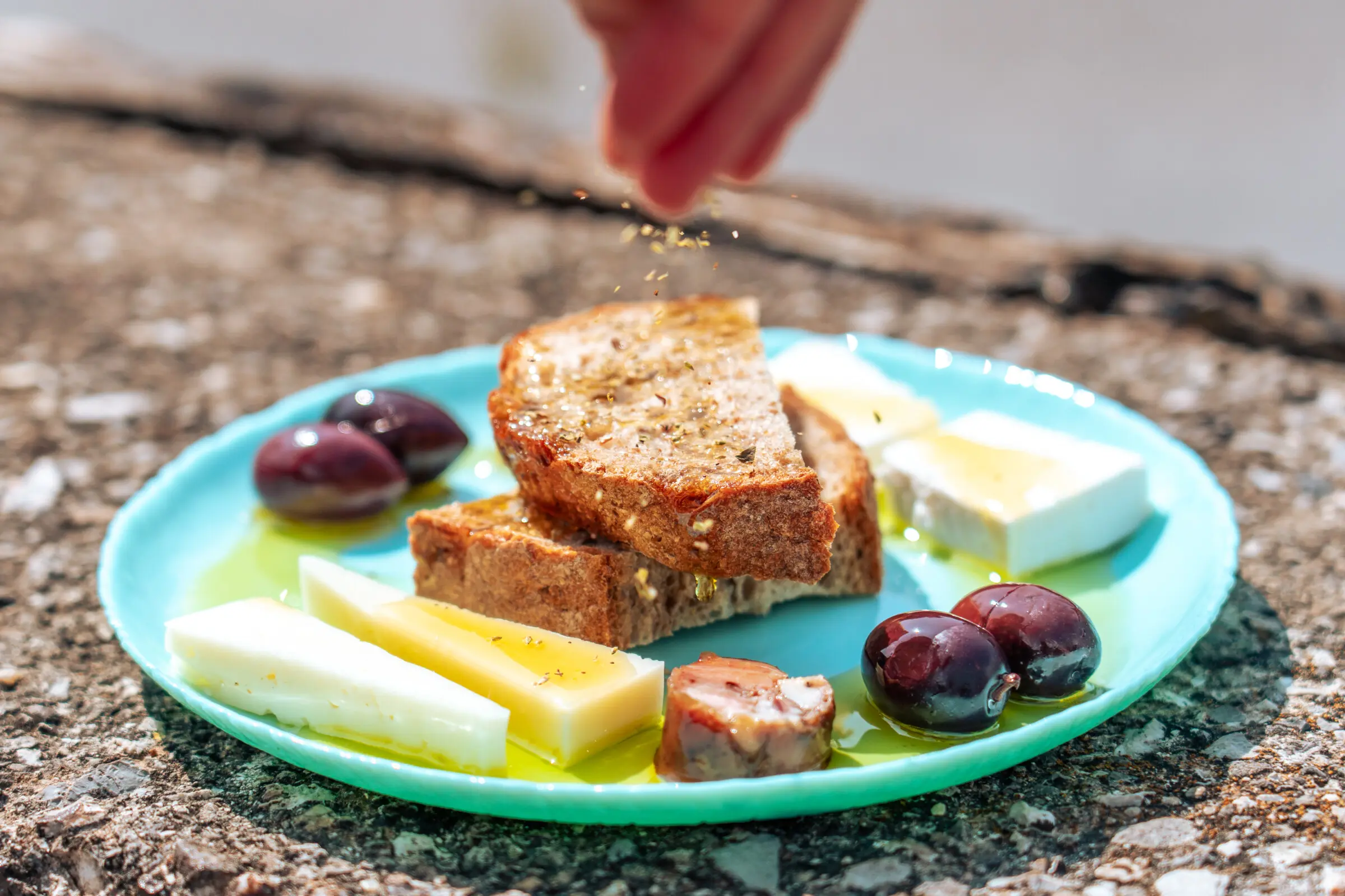 This is a close-up of a plate with bread, cheese, olives on it. A hand sprinkles the bread with dried oregano.