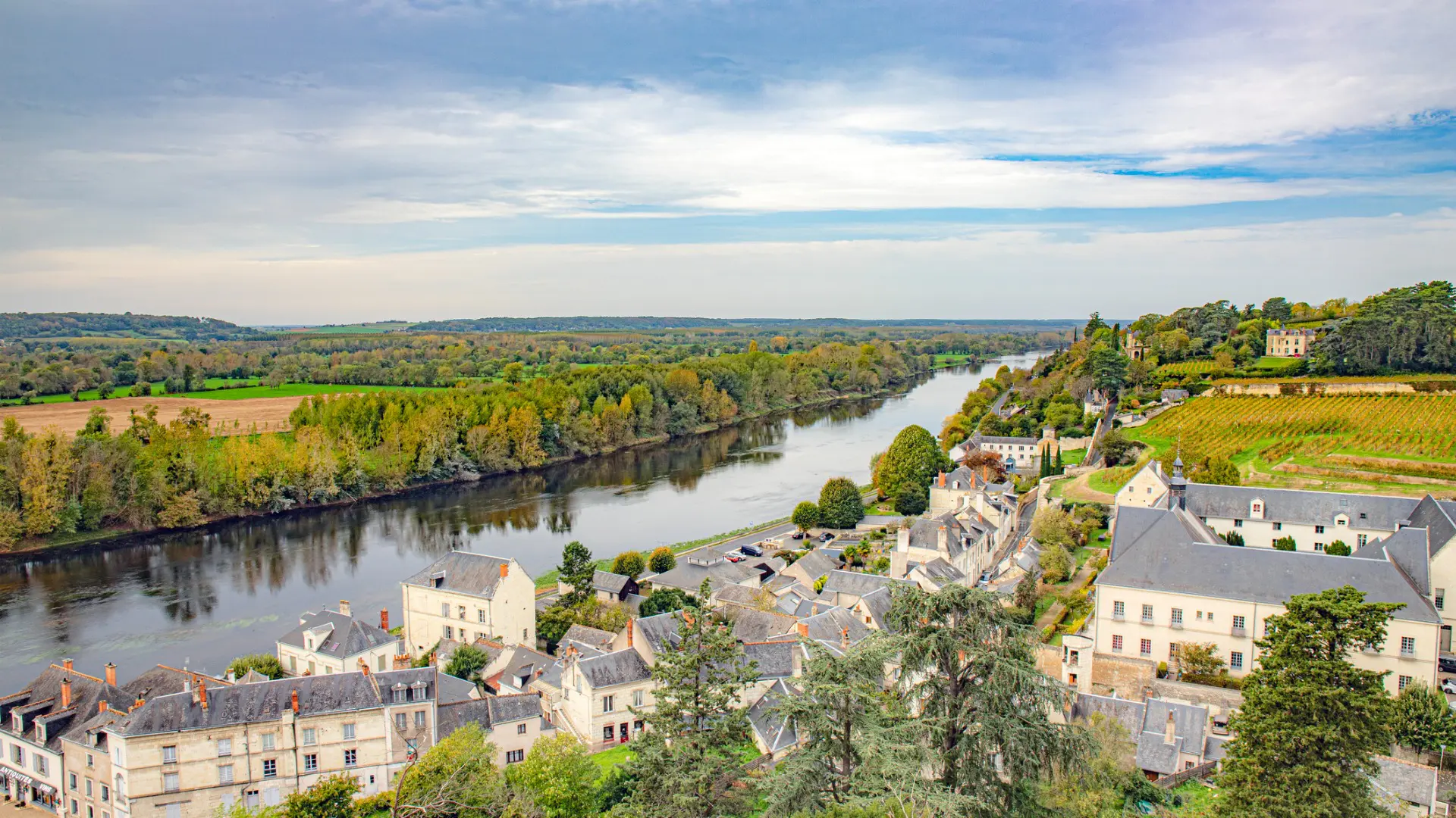 This image shows a river flowing through a valley. One bank is covered with lush vegetation. On the other, a quaint town with white and grey buildings is built.