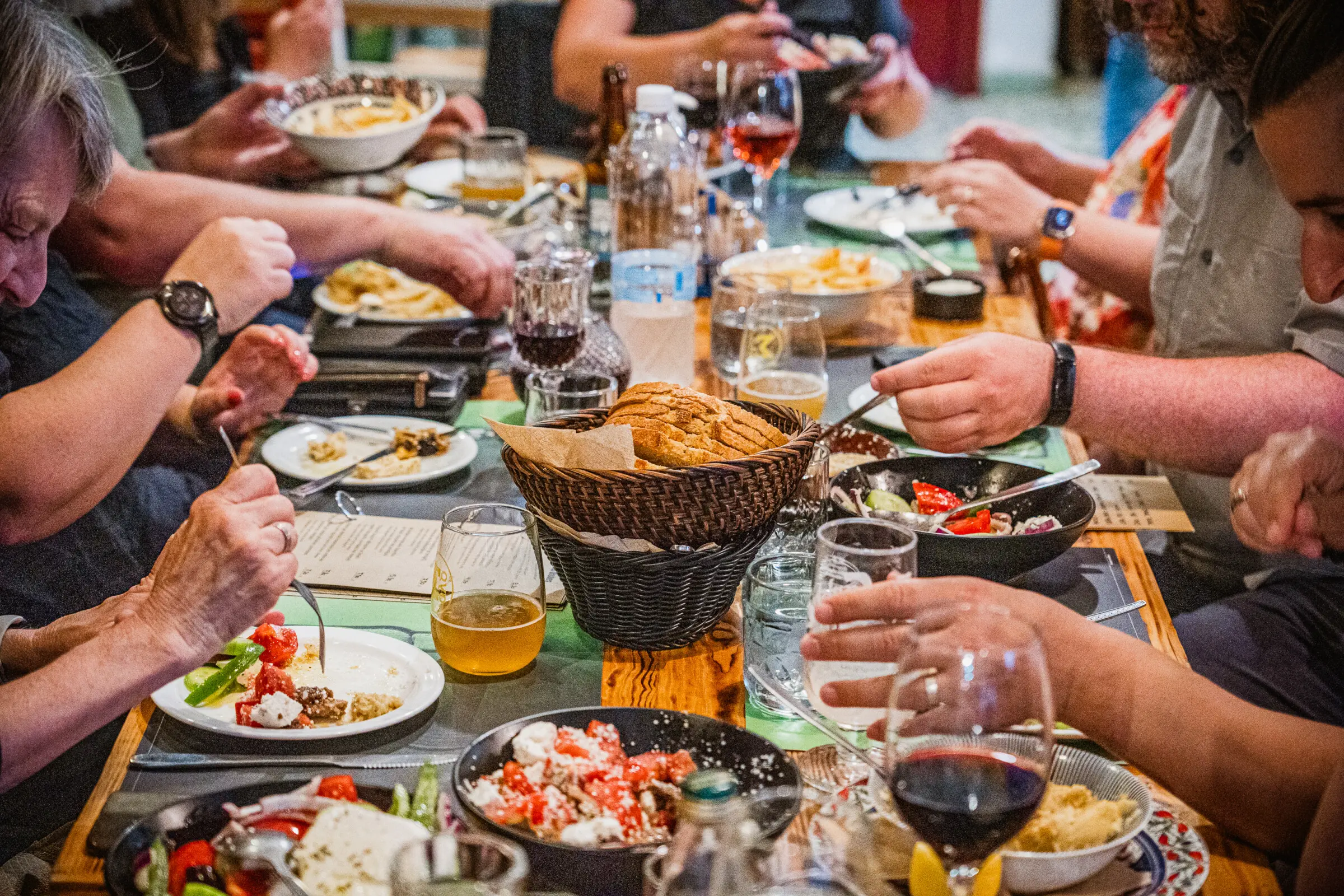 This image shows a long table full of Greek dishes and the hands of several people eating.