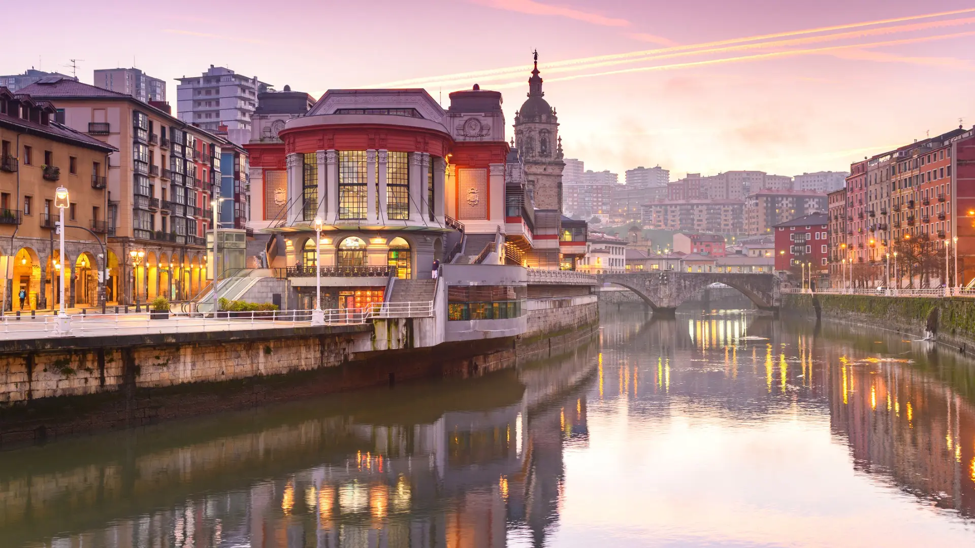 Quaint buildings on both sides of a calm river under a pink sunrise sky.