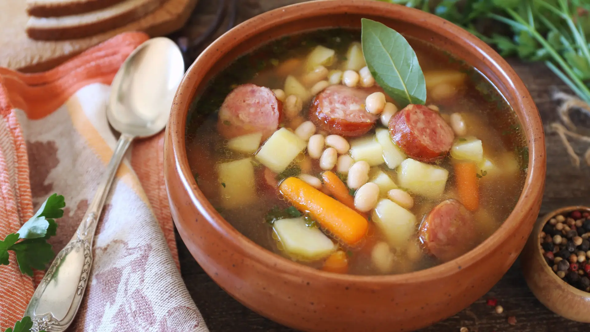 This image shows a bowl of Caldo Gallego and a spoon next to it. In the soup, there are pieces of sausage, white beans, carrots, and other vegetables.