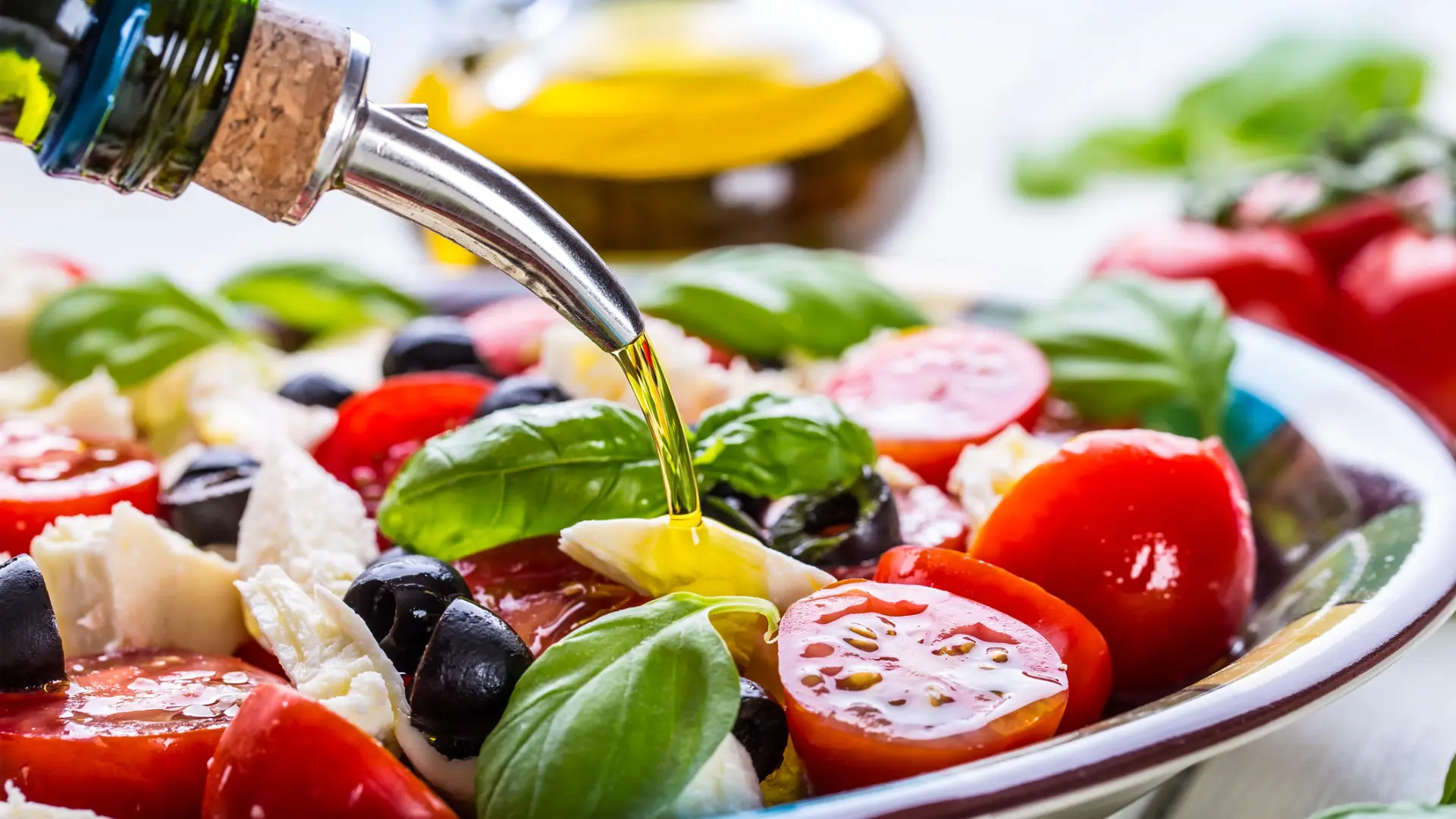 A close up of a Caprese salad with tomatoes, mozzarella, black olives, and basil leaves as olive oil is poured on top.