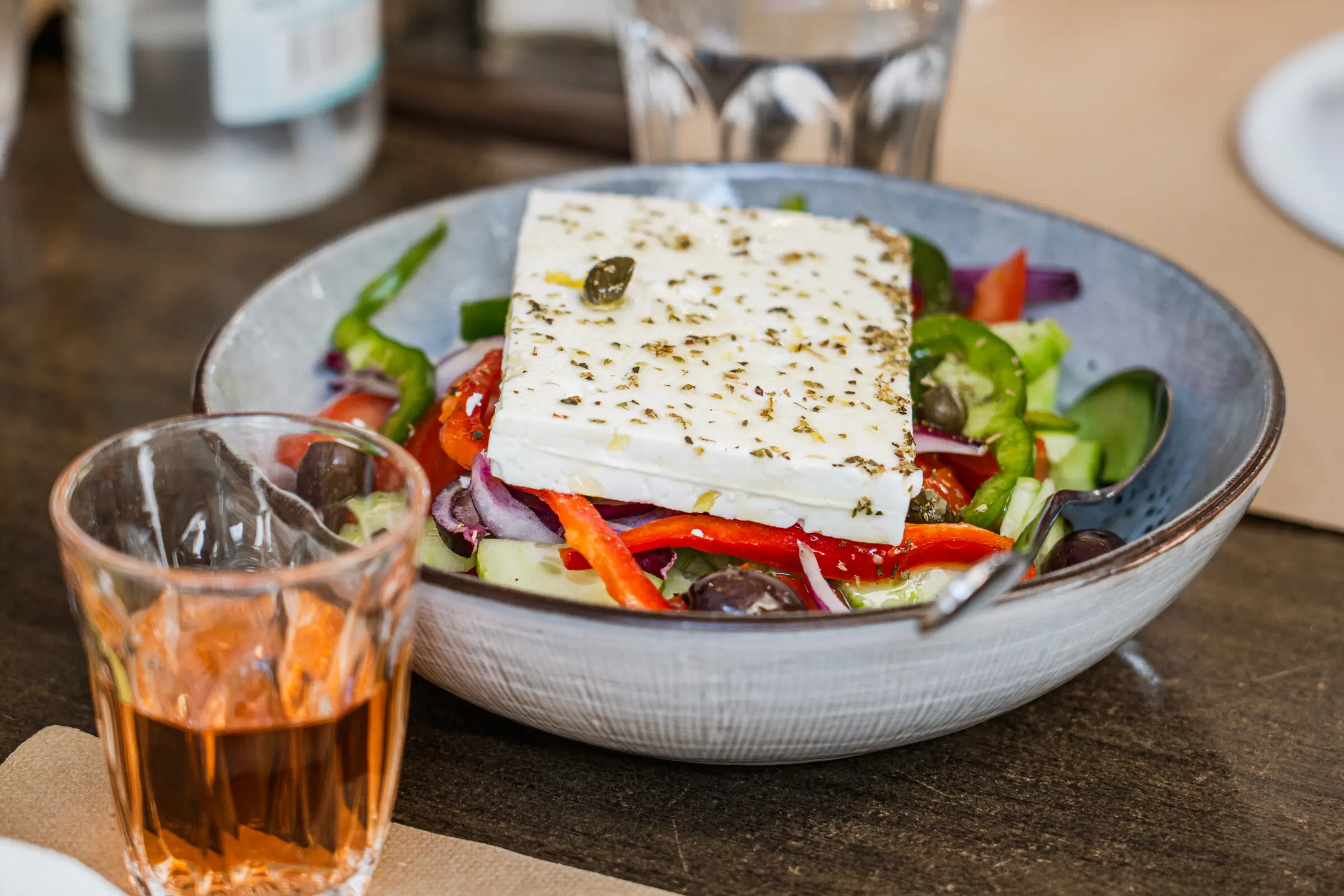 A close up of a Greek salad with cucumbers, red bell peppers, green bell peppers, olives, onions, and a large piece of feta cheese on top. Next to the bowl, a small glass of rose wine.