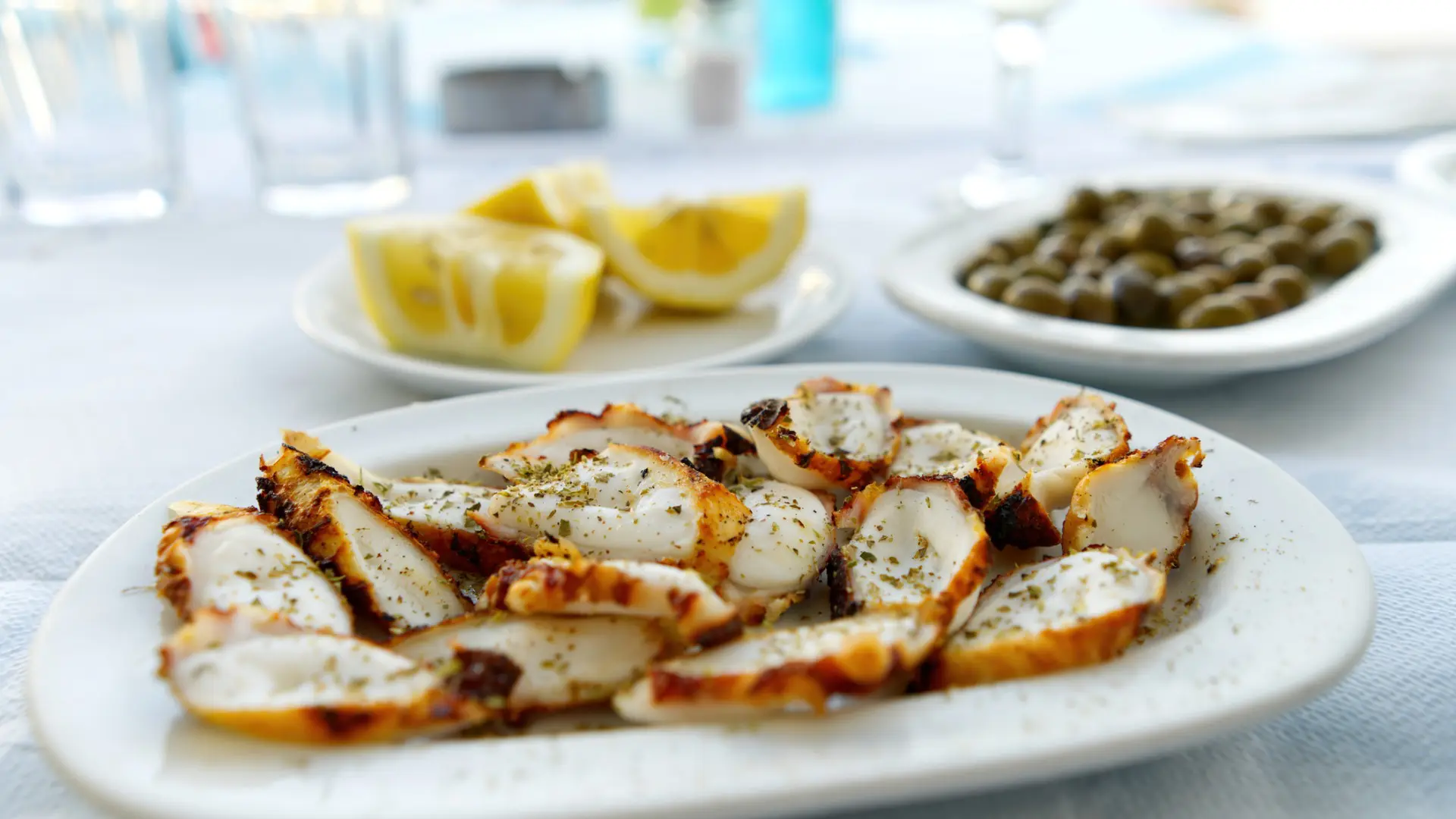 In the foreground, a white plate with pieces of grilled octopus. In the background, a small plate with lemon wedges and another with olives.