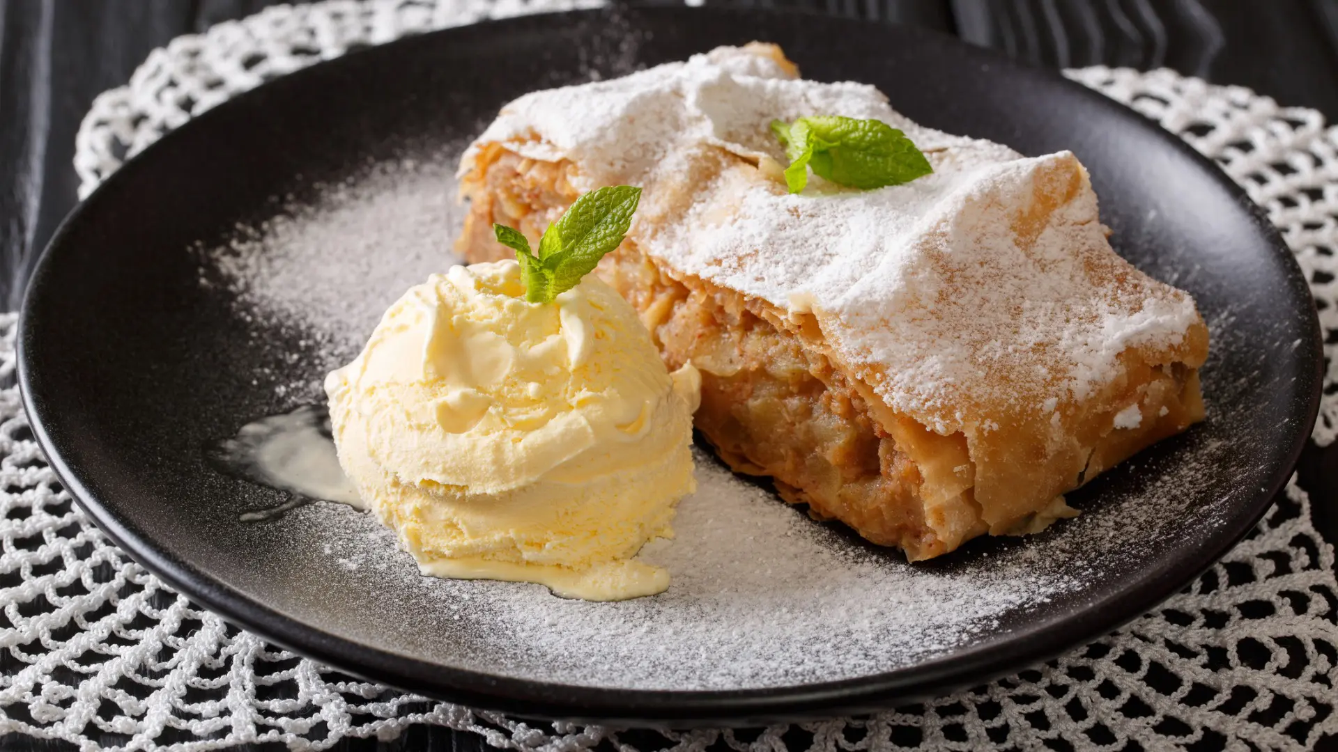 A black plate with a slice of apple strudel topped with powdered sugar and paired with vanilla ice cream.