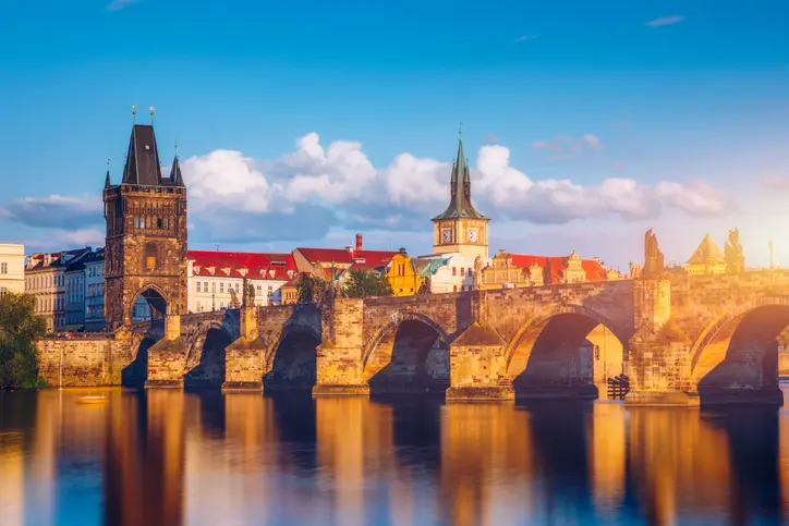 A beautiful view of the historic Charles Bridge in Prague, Czech Republic, captured during the golden hour. The bridge, with its stone arches and iconic Gothic towers, spans the calm Vltava River, reflecting the warm hues of the setting sun. The picturesque scene is complemented by the classic architecture of the surrounding buildings, bathed in soft sunlight under a clear blue sky with scattered clouds. This image highlights the timeless charm and serene beauty of one of Prague's most famous landmarks.
