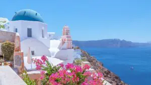 Iconic view of the blue-domed church in Oia in Santorini, with the caldera in the background and bright pink flowers in the foreground.