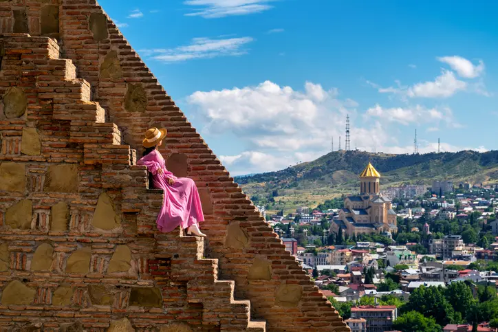A captivating image of a woman in a flowing pink dress and straw hat sitting on the ancient stone steps of a historic fortress, overlooking the cityscape of Tbilisi, Georgia. The composition beautifully captures the contrast between the rugged, time-worn bricks of the fortress and the vibrant, modern city below. The iconic Holy Trinity Cathedral, with its golden dome, stands out in the distance against the backdrop of rolling hills and a bright blue sky dotted with fluffy clouds. This scene blends cultural heritage with the scenic beauty of Tbilisi.