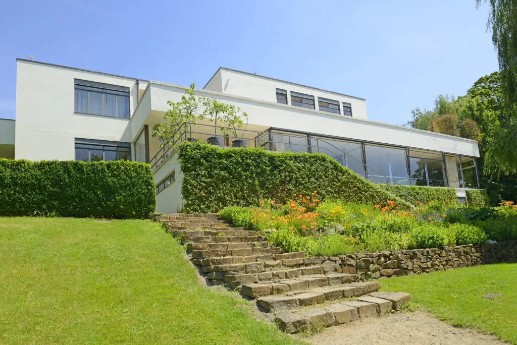 A photograph of the Villa Tugendhat, a modernist architectural masterpiece located in Brno, Czech Republic. The building features clean lines and a minimalist design with large glass windows, surrounded by well-manicured gardens. A stone staircase leads up to the house, bordered by lush greenery and colorful flowers, blending the structure seamlessly into its natural surroundings.