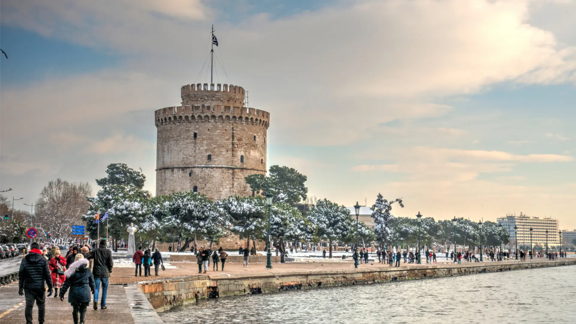 The White Tower in Thessaloniki. There are some trees capped with snow and many people walking along the promenade.