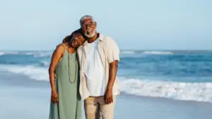 This image shows a smiling older couple standing close together on a beach. The man is wearing a light-colored shirt and pants, while the woman is in a flowing green dress with long beaded necklaces. The woman leans affectionately on the man's shoulder as they pose together in front of the ocean, with gentle waves in the background under a clear sky. The mood is warm and relaxed, capturing a peaceful and joyful moment by the sea.