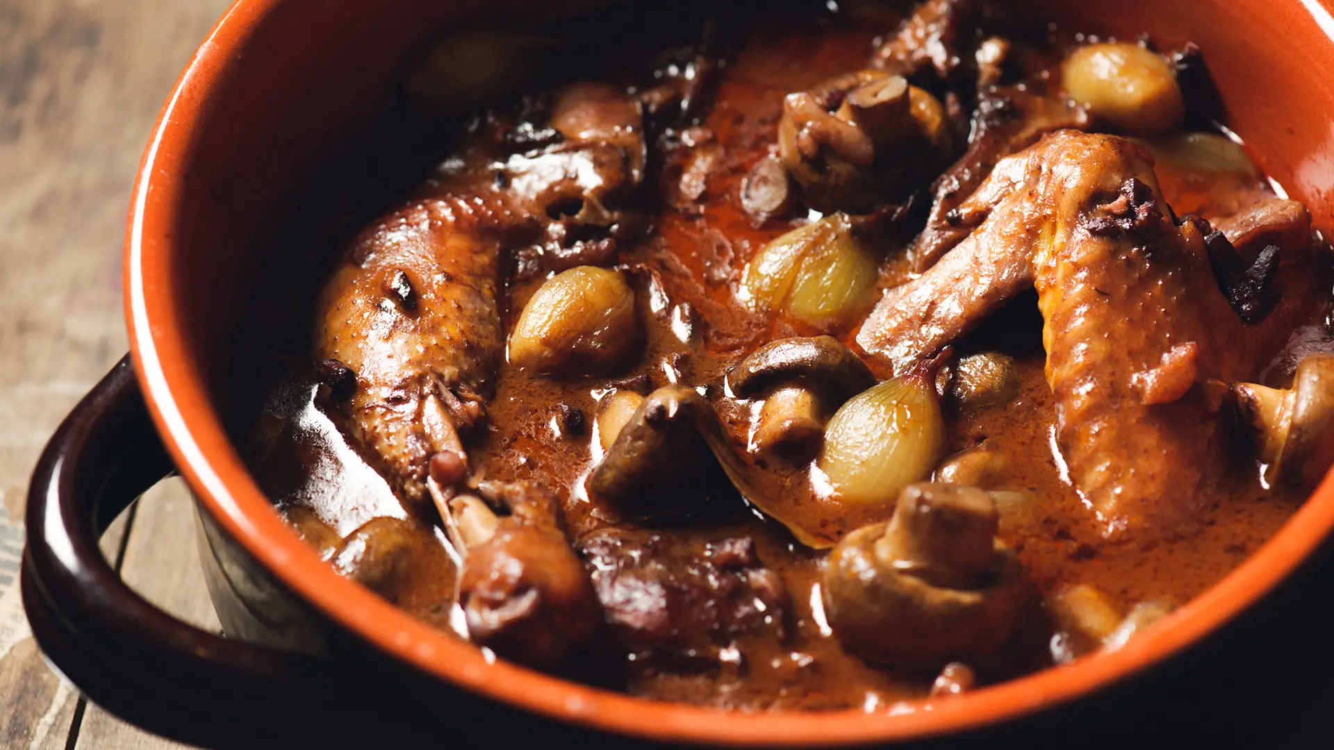 A close up of a clay pot with Coq au Vin or chicken stew with mushrooms and onions in a tomato sauce.