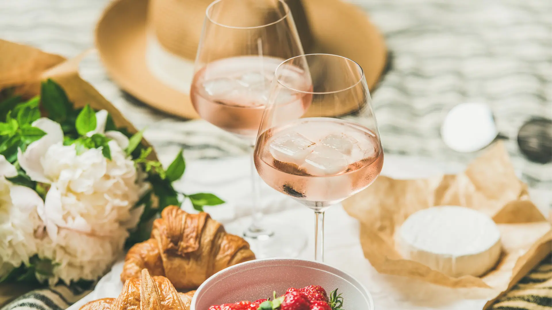 This image shows two glasses of Provencal rose wine, brie cheese, two croissants, a bowl with strawberries, white flowers, and a hat in the background.