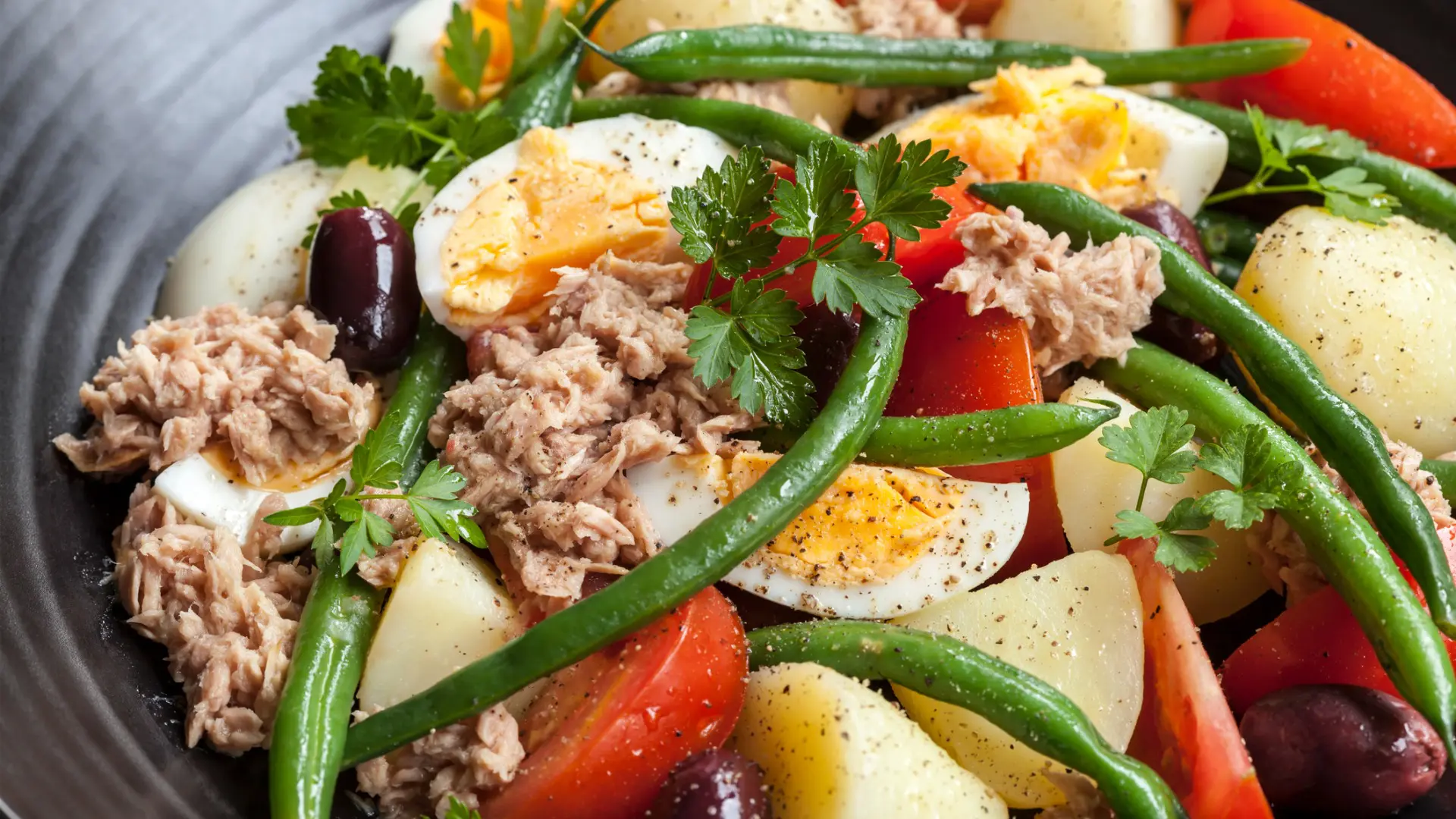A close up of a salad with tuna fish, hard-boiled eggs, green beans, tomatoes, boiled potatoes, and parsley.