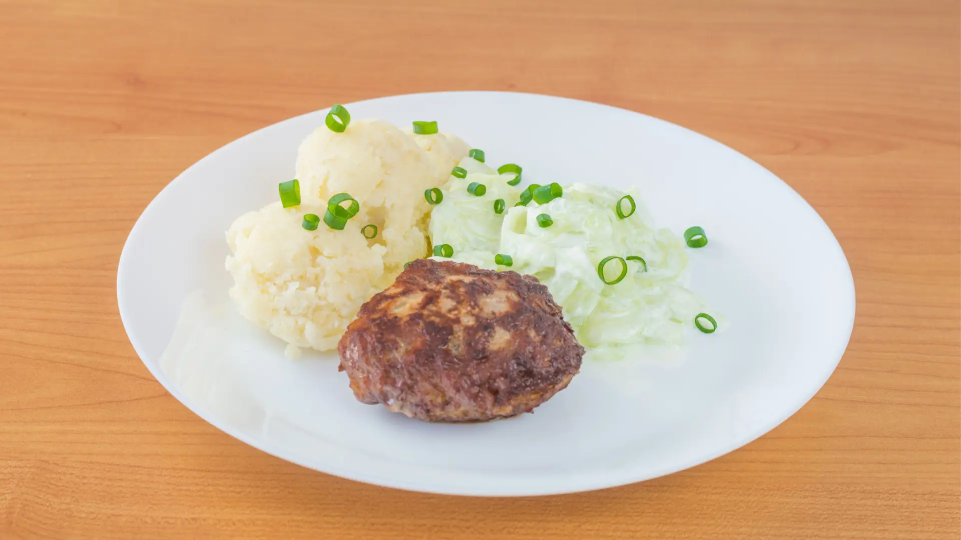 A white plate with a meat patty, mashed potatoes, and a salad made of sliced cucumbers.