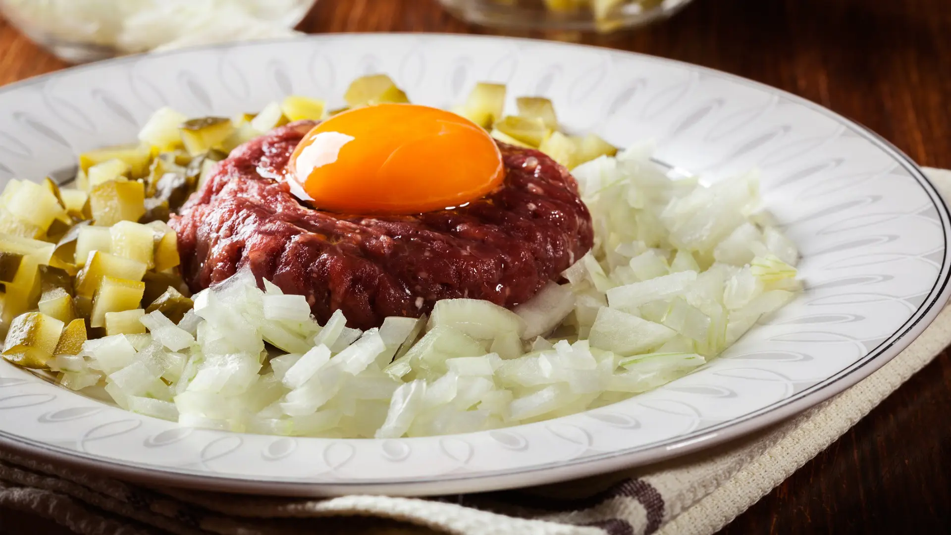 A white plate with a raw beef patty sitting on chopped onions and pickles and topped with a raw egg yolk.