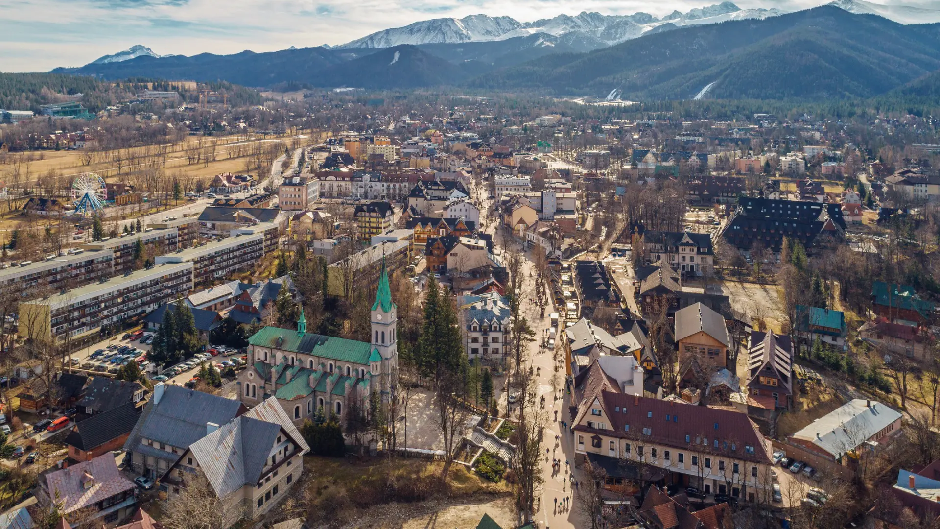 Aerial view of the town of Zakopane with tall mountains in the background. There are both traditional buildings and austere blocks of flats in the town.