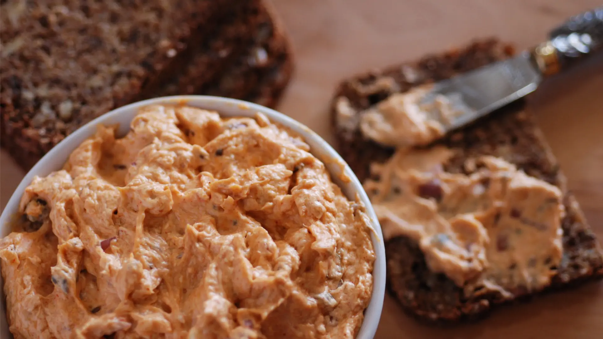 A white bowl filled with an orange dip in the foreground. A knife spreading some of this dip on a slice of rye bread in the background.