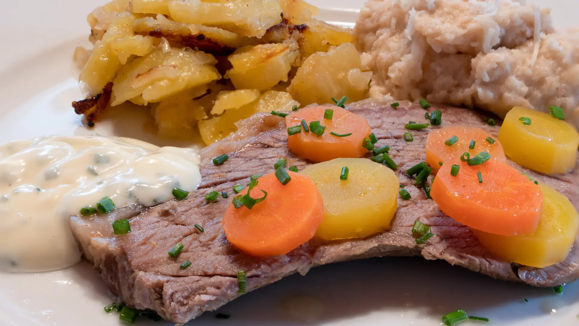 A piece of meat with carrots on top, potatoes and a white sauce as sides.