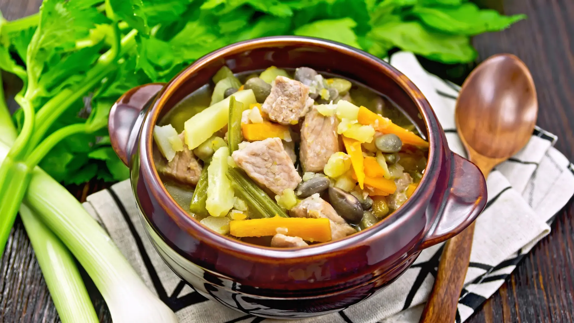 Eintopf soup made from pork, celery, beans, carrots, and potatoes with leek in a clay bowl on a napkin on a dark wooden board background.