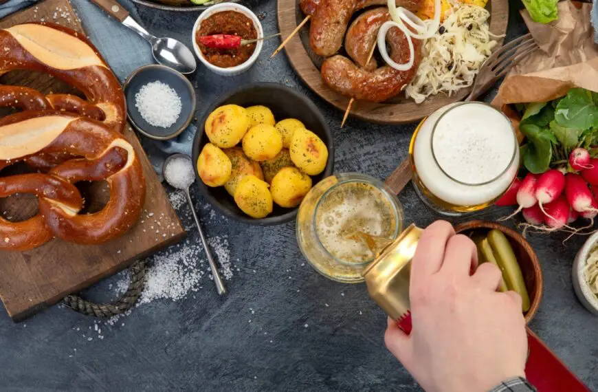 Pretzels, potatoes, sausages and vegetables on a dark surface. A hand is pouring beer from a can into a glass.