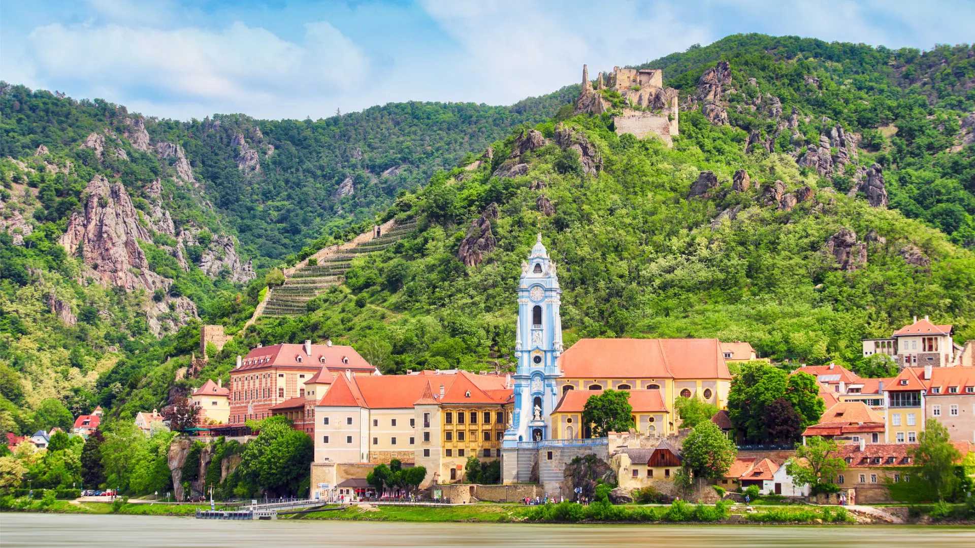 A picturesque village by the river with vineyards and a hilltop fortress in the background.
