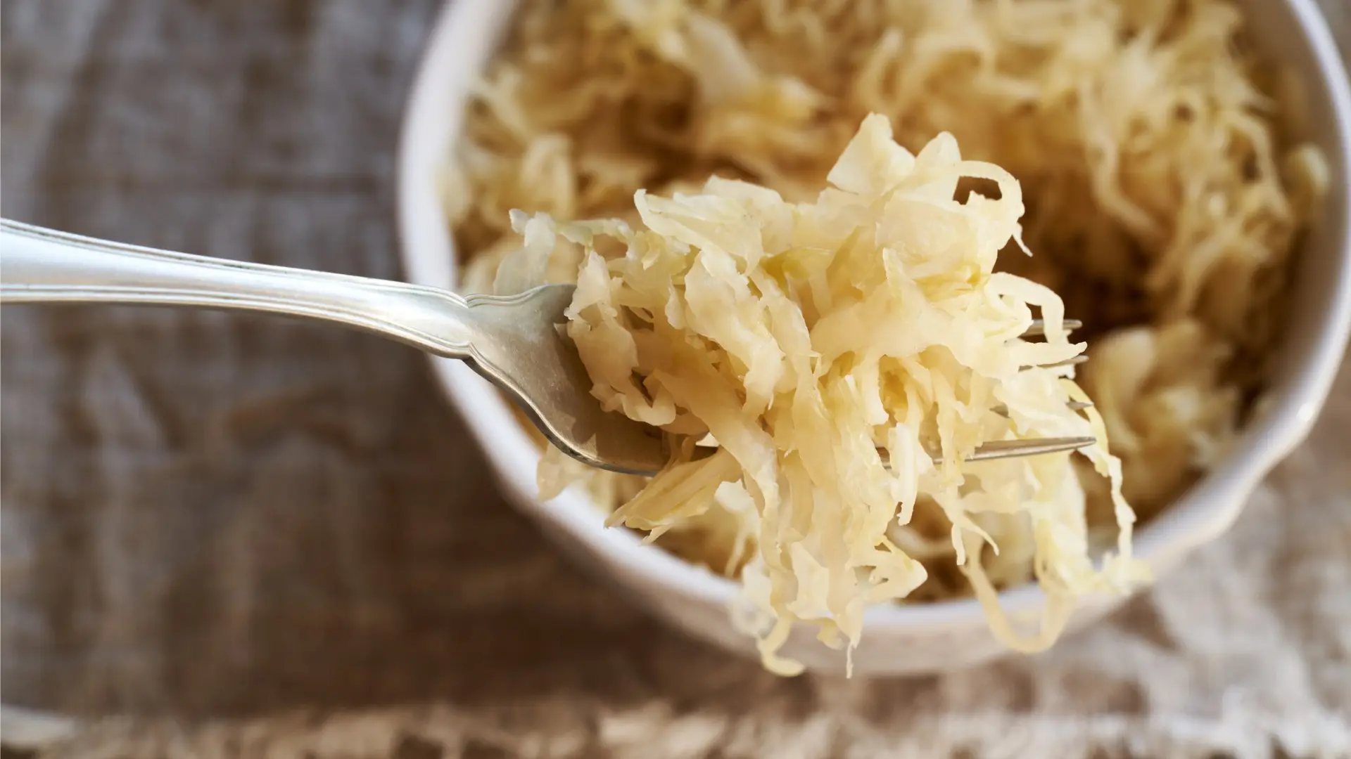 A close up of a fork with Sauerkraut and a white bowl filled with Sauerkraut in the background.