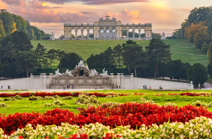A panoramic view of the Gloriette overlooking the Schonbrunn Gardens under a glorious sunset sky. There are flowers of vivid colors in the foreground.