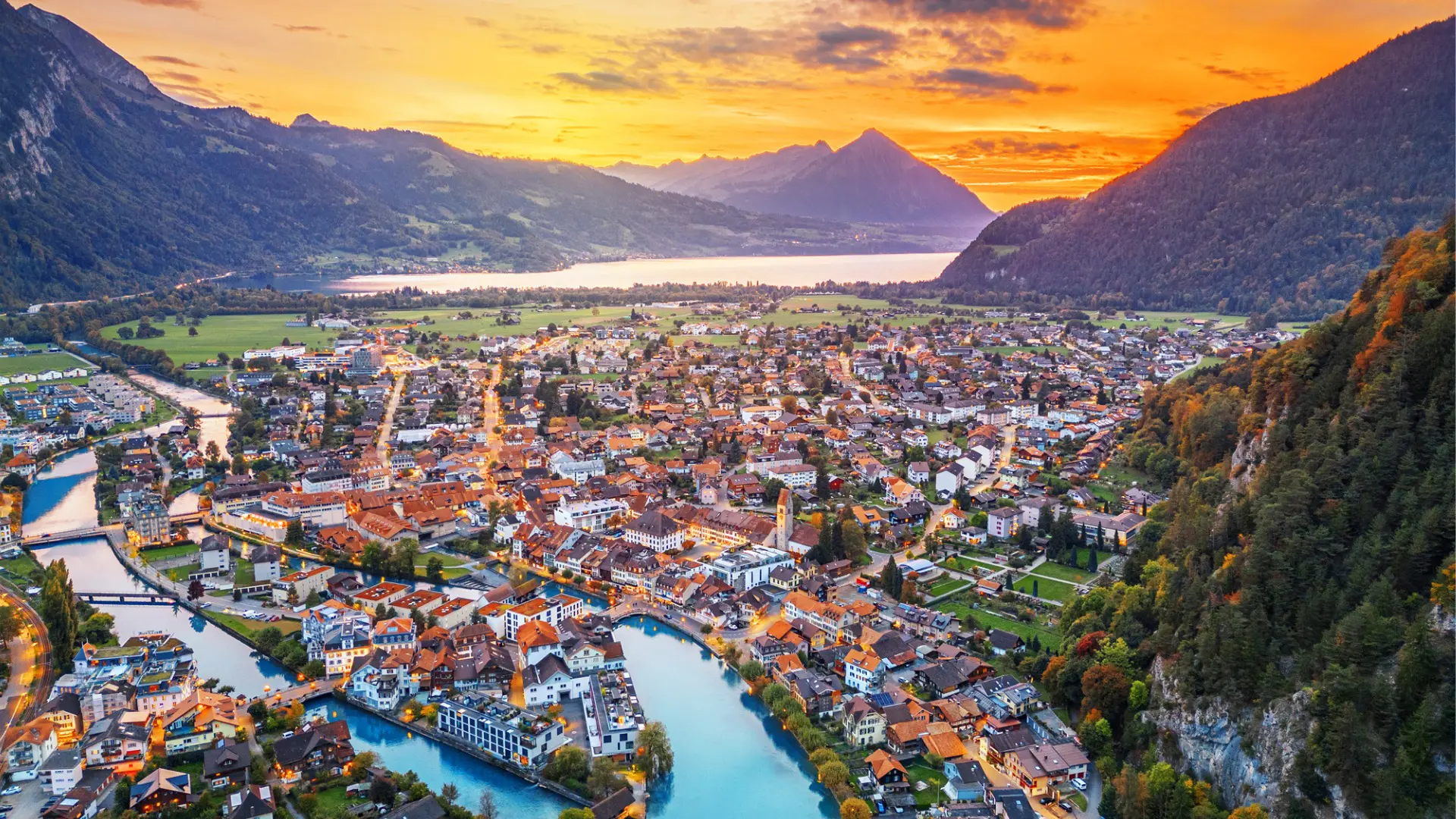 An aerial view of Interlaken, a beautiful town with a river. There are mountains and a lake in the distance, under an orange sunset sky.
