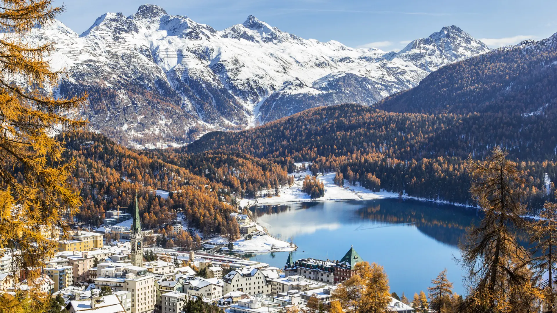 Tall snow-capped mountains and a small town by a lake.