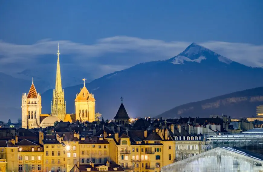 Geneva's illuminated St. Pierre Cathedral at dusk with a snow-capped mountain in the background and a row of beautiful buildings in the foreground.