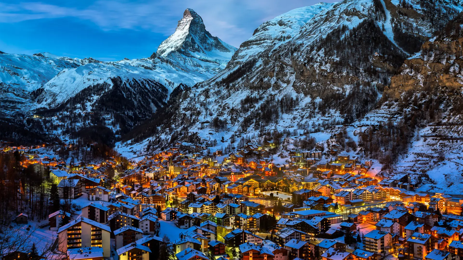 An illuminated town nestled in a valley dominated by a high snow-covered peak.