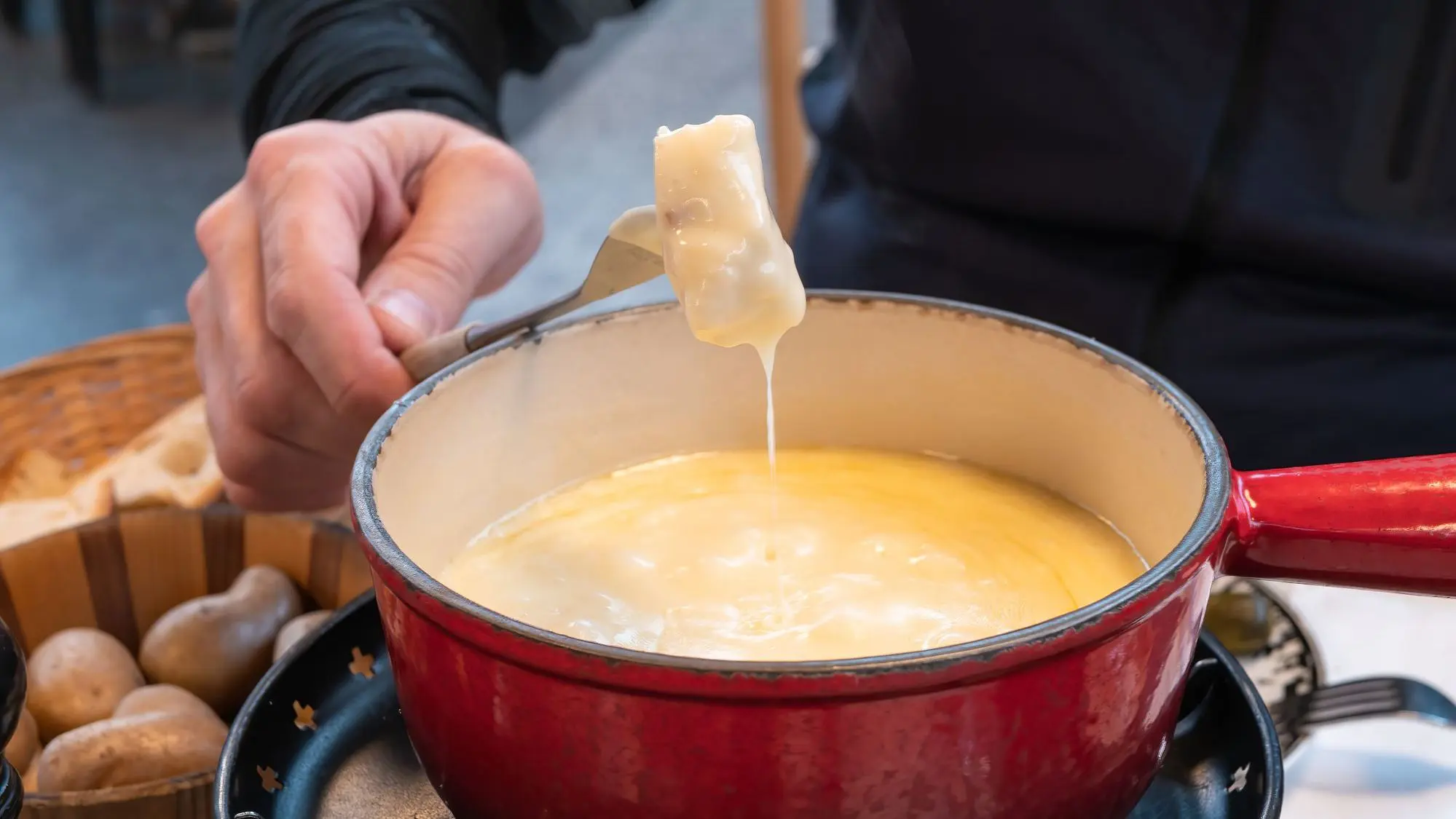 A man's hand holds a fork with a piece of bread that has been dipped in a cheese fondue pot.