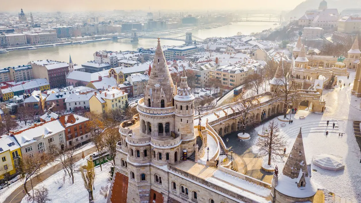 Aerial view of the snowy Fisherman's Bastion with the Szechenyi Chain Bridge and St. Stephen's Basilica in the background on a snowy winter morning.