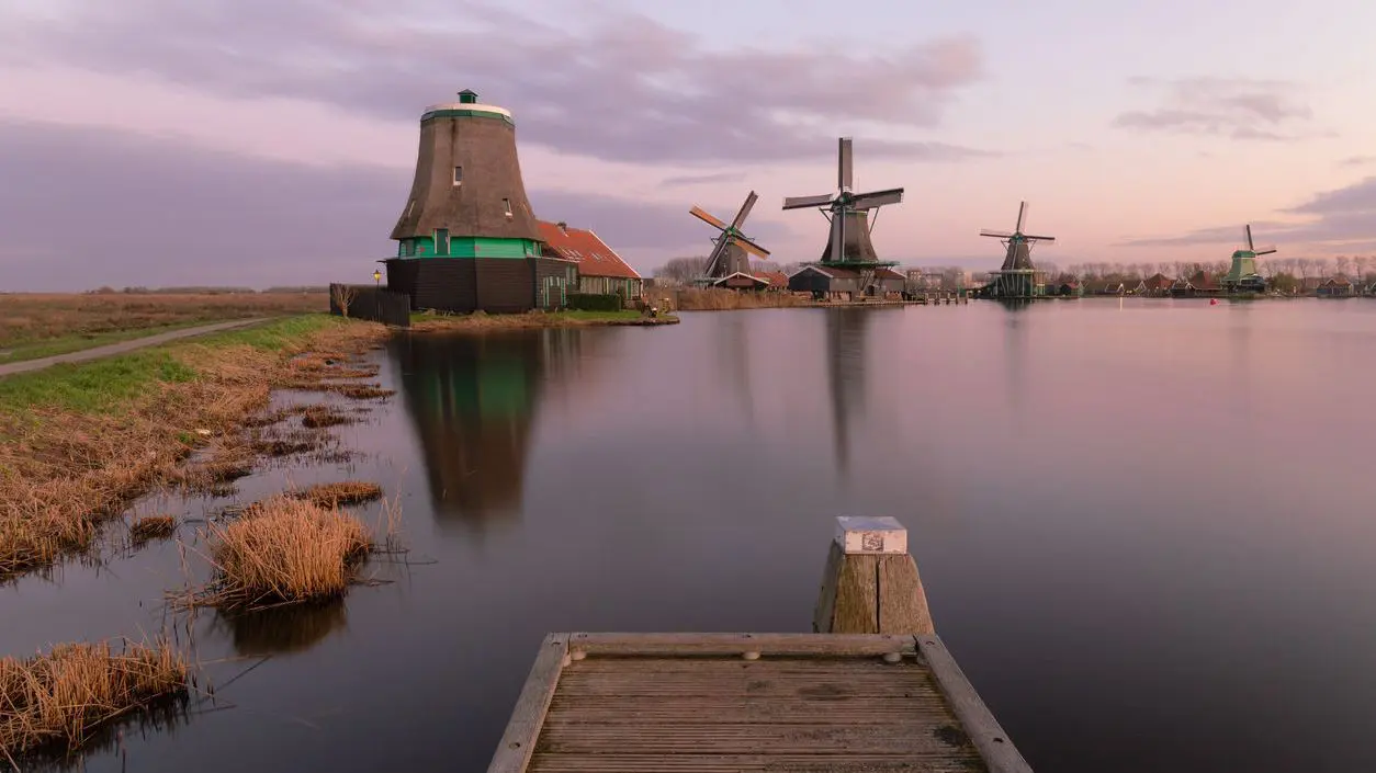 A dreamy landscape of wooden windmills by the water under a lilac sky.