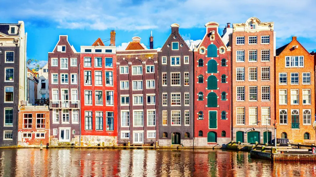 A row of typical Amsterdam buildings of various colors, lining a canal.