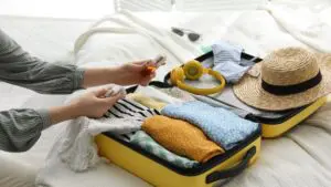 A small yellow suitcase open on a bed with clothes folded inside and a straw hat and yellow headphones on top of them. A woman's hand is holding a perfume over the suitcase.